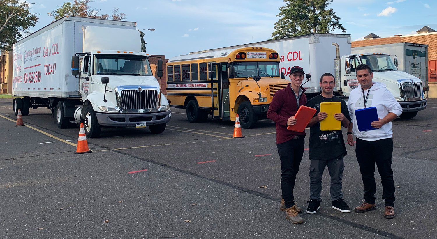 Three people with clipboards standing in front of tractor-trailer, bus, and box truck