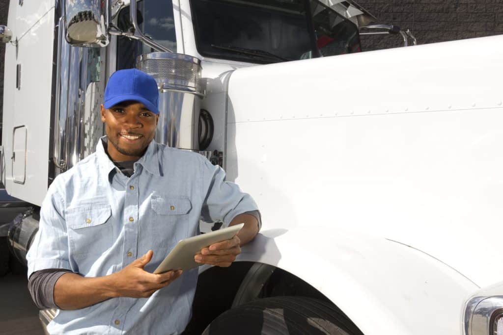 Smiling driver leaning against big rig truck and holding a tablet
