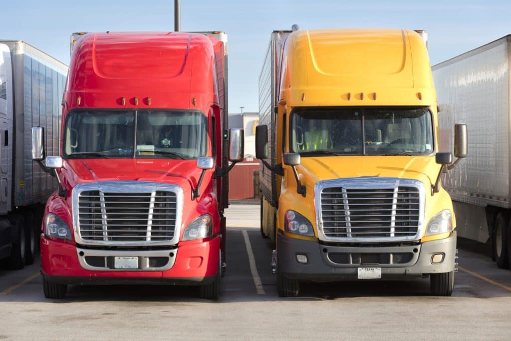 Two tractor-trailers, one with red cab, the other with yellow cab, parked in parking lot